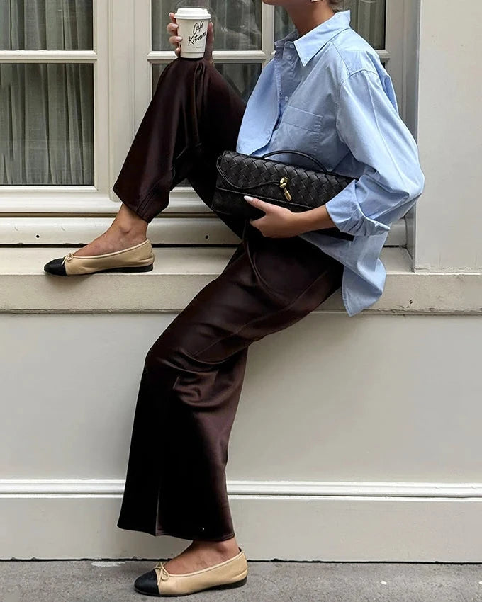 Person sitting on steps wearing a light blue shirt, brown pants, and beige flats, holding a coffee cup and black handbag.