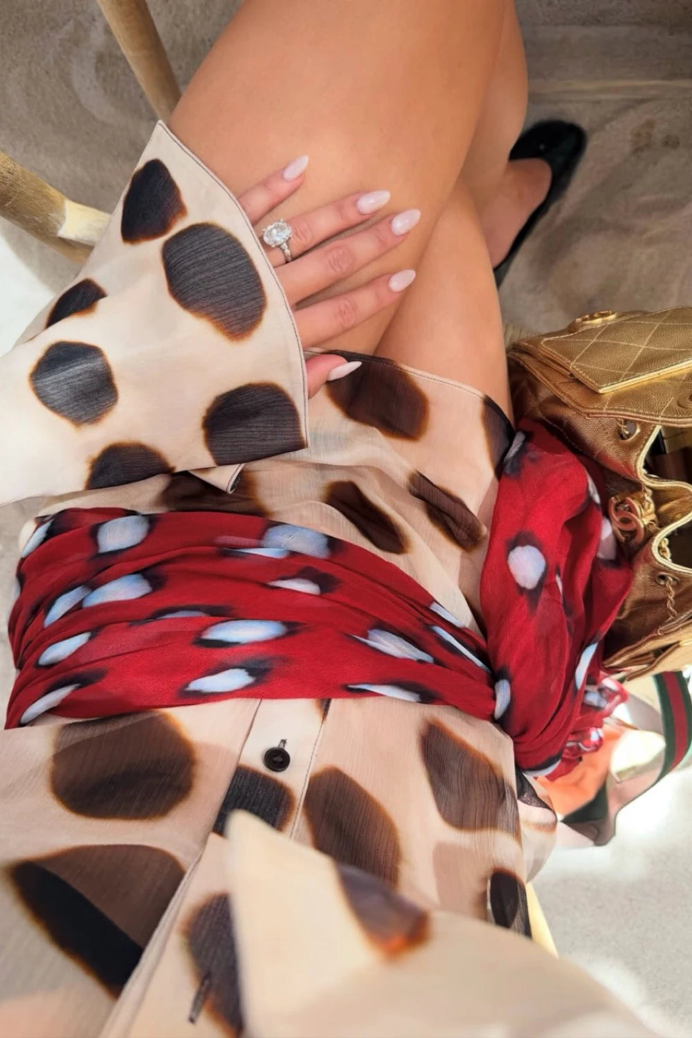 A close-up lifestyle shot of a person wearing a beige sheer blouse with a brown and black leopard print, a red patterned scarf, and black flats, with a gold handbag nearby.