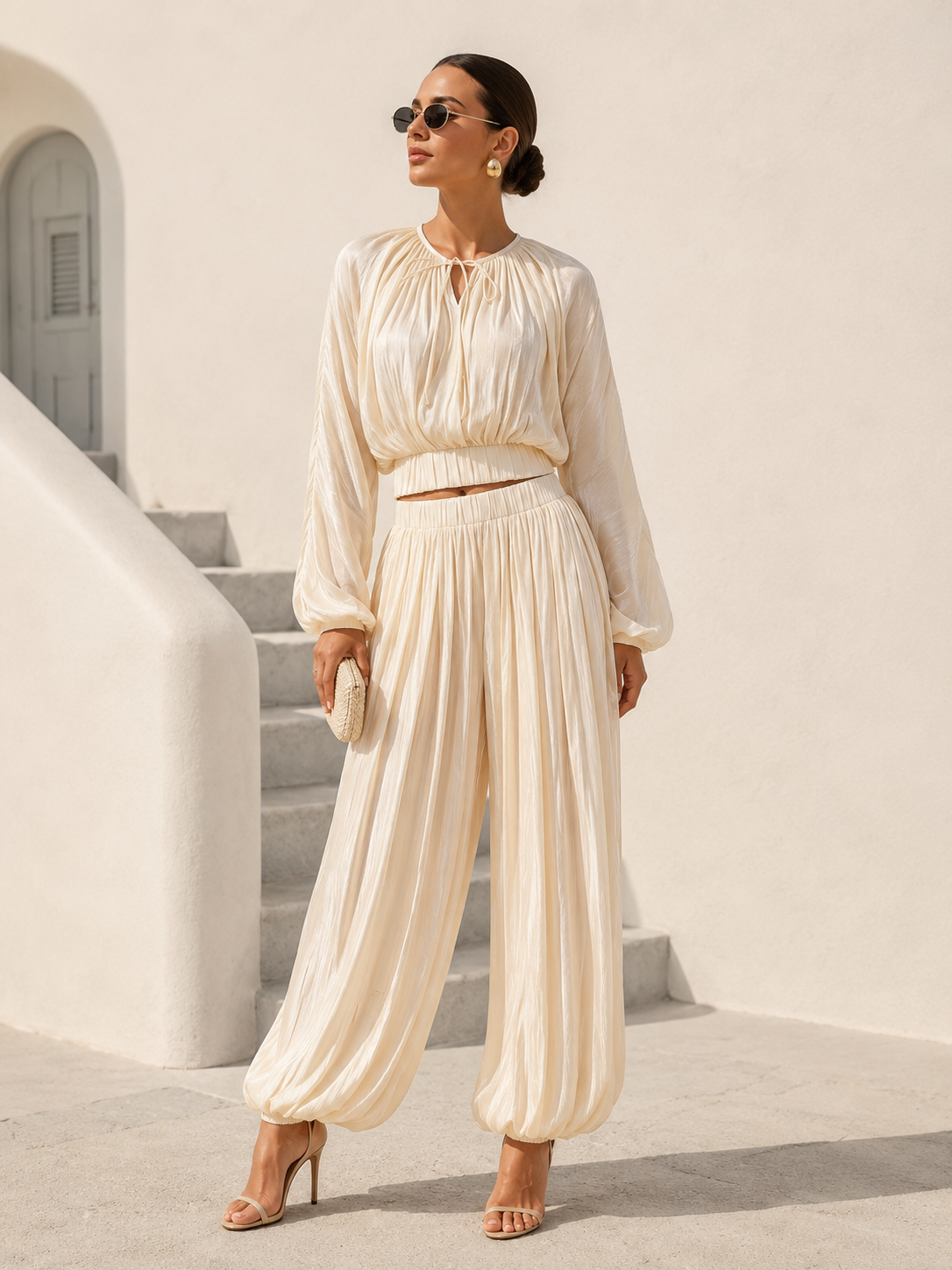 A woman models a cream-colored two-piece set consisting of a long-sleeve tie-front blouse and matching pleated wide-leg pants, styled with sunglasses, a small clutch, and high-heeled sandals against a white stucco background.