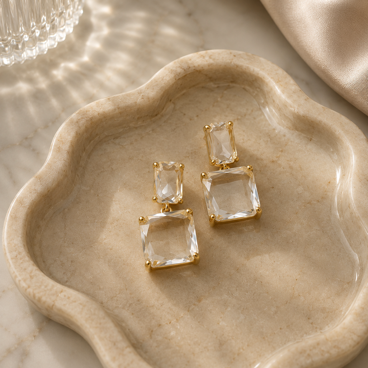 A pair of gold-tone drop earrings with clear rectangular gemstones, displayed on a beige scalloped tray over a marble surface with soft satin fabric in the background.