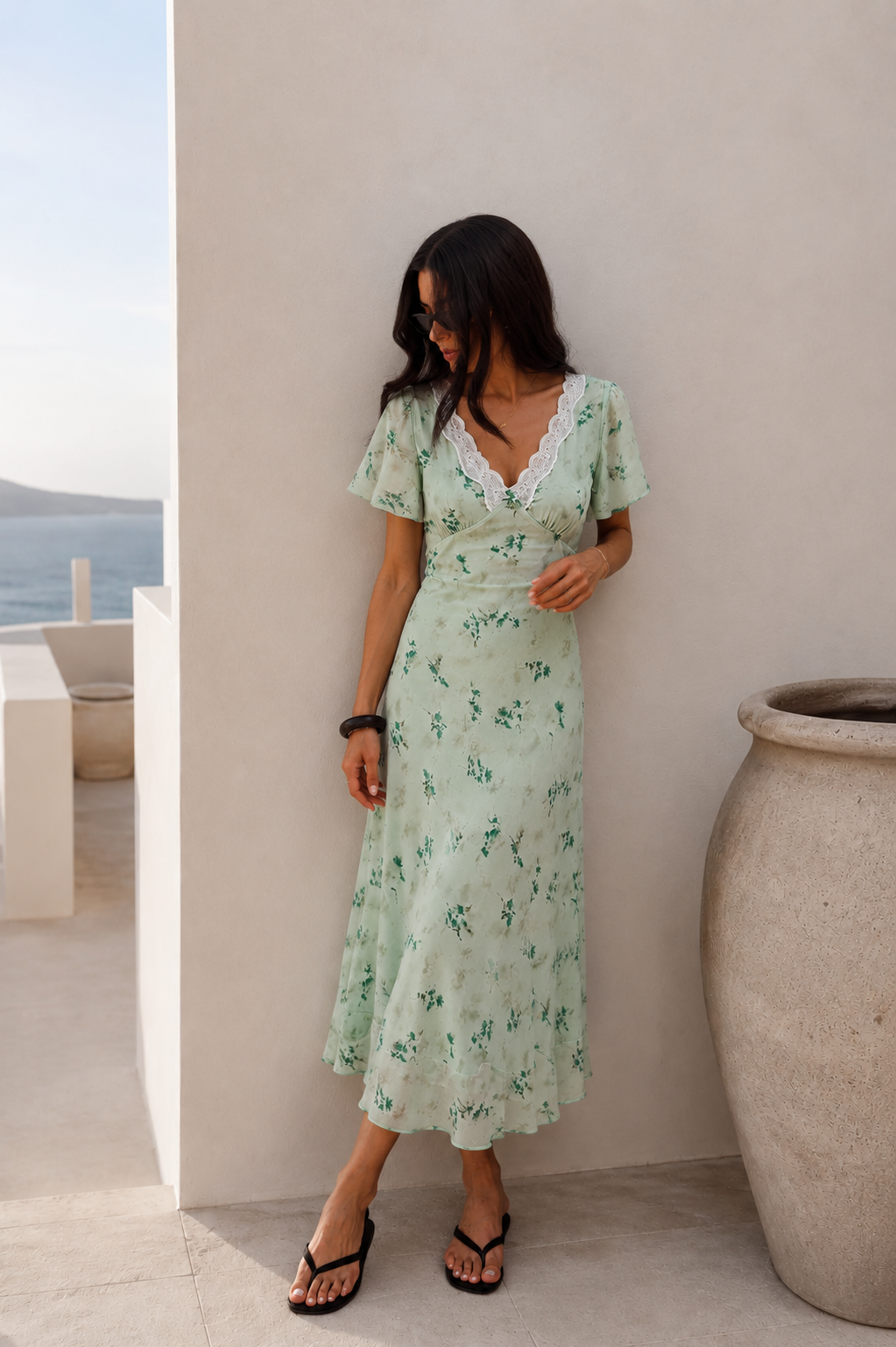A woman wearing a light green floral midi dress with white lace trim stands on a sunlit balcony next to a large beige planter pot.
