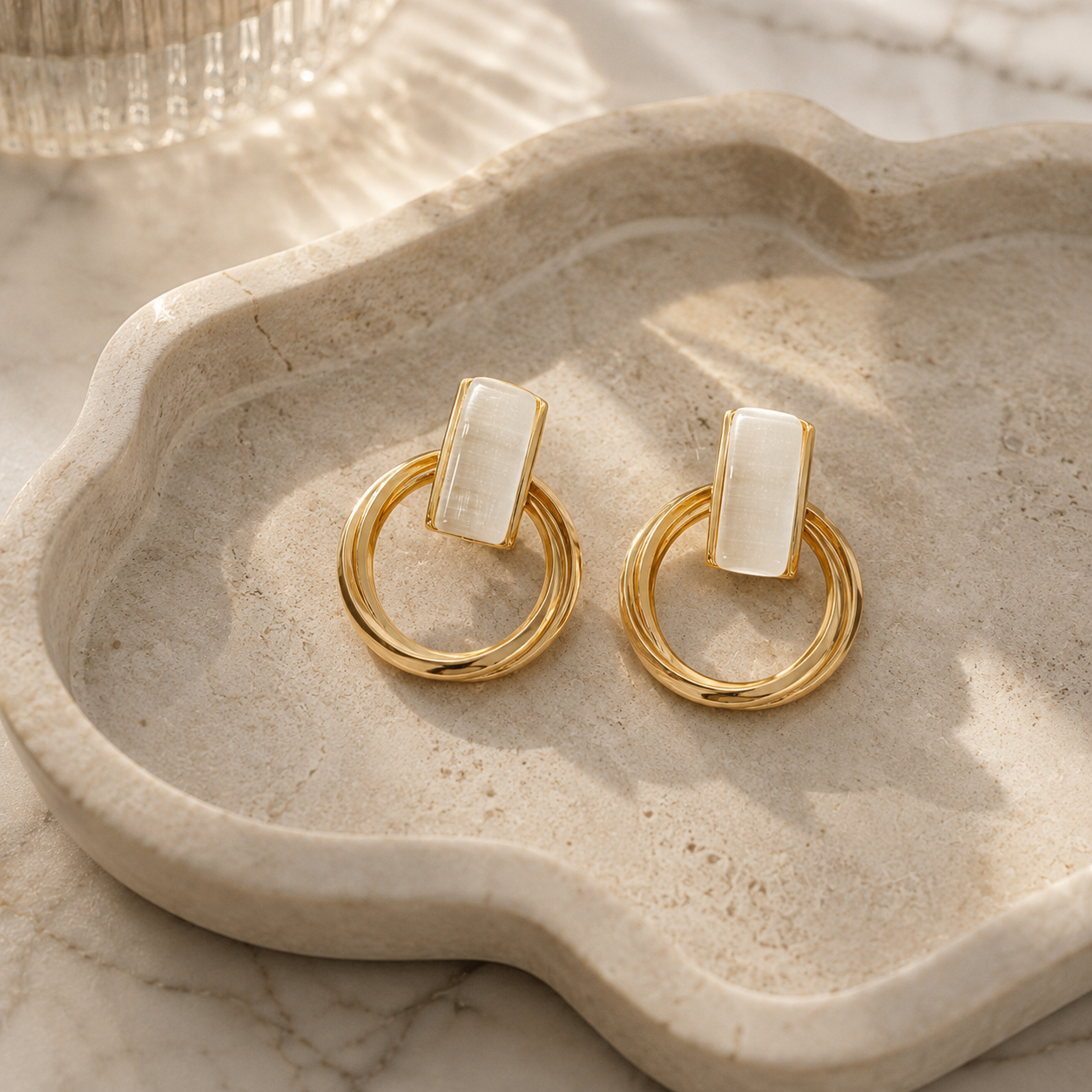 A pair of gold hoop earrings with rectangular white inlay stones, displayed on a beige stone tray.