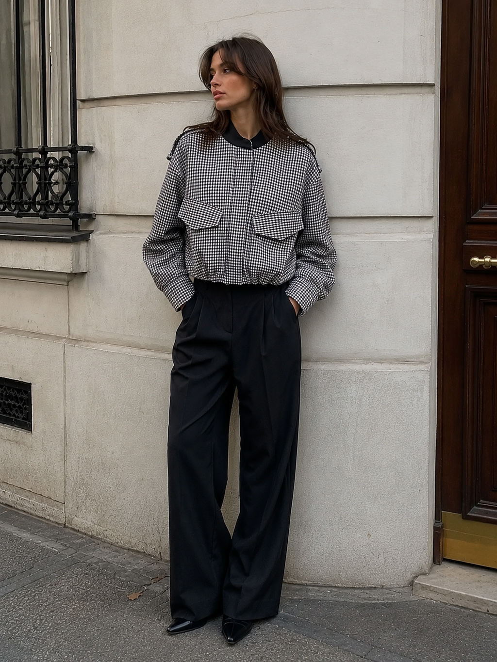 A woman stands against a light stone building wall wearing a black and white gingham cropped jacket with a black collar and flap pockets, paired with black wide-leg trousers.