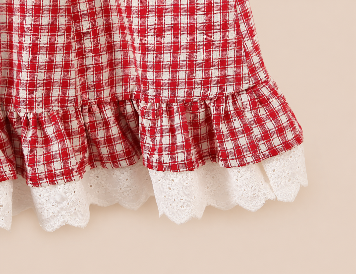 Close-up of a red and white plaid fabric with ruffled edges and white eyelet lace trim, shown against a plain beige background.