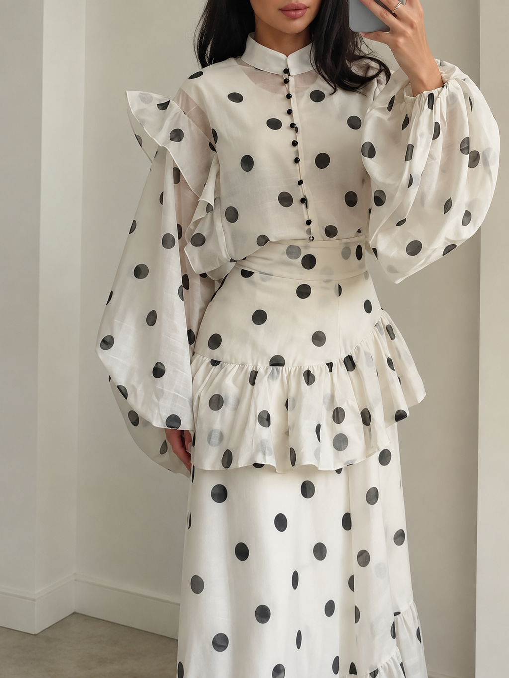 A woman wearing a white long-sleeve blouse and matching skirt set with black polka dots, featuring ruffled details and a high collar, photographed indoors against a light neutral wall.