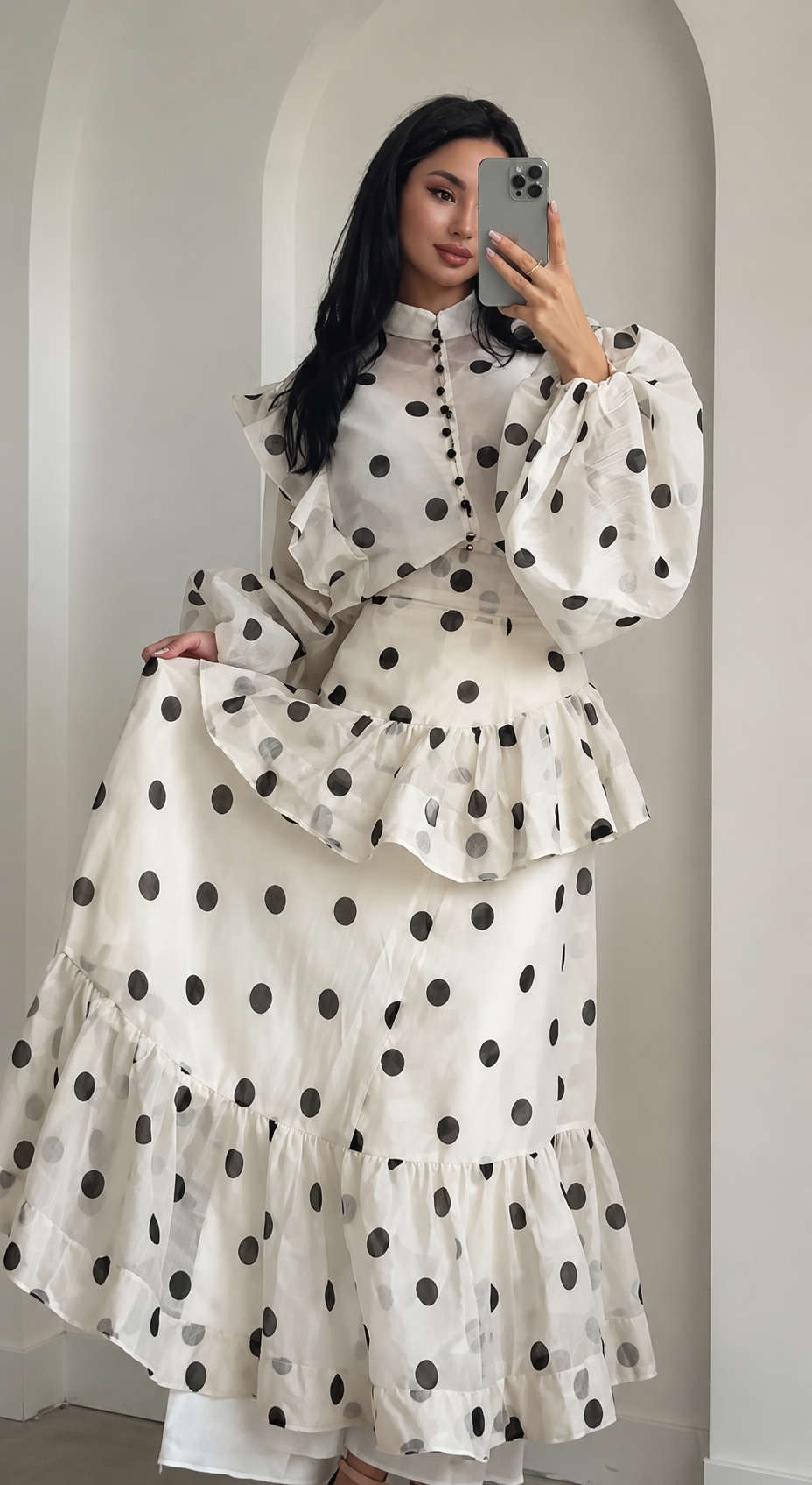 A woman takes a mirror selfie wearing a long-sleeved white dress with black polka dots, featuring a high ruffled collar, a ruffled waist, and a tiered skirt.