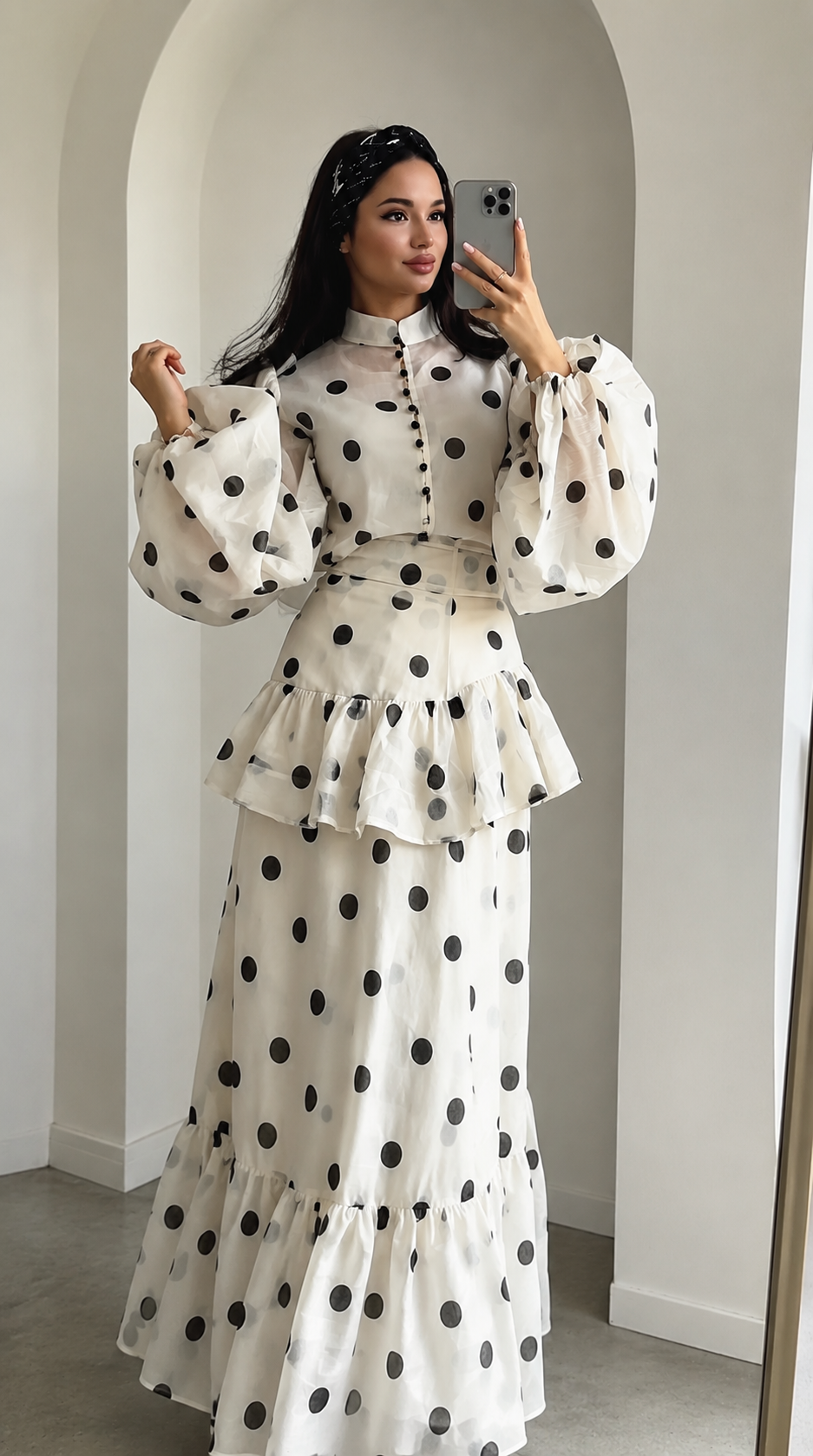 A woman takes a mirror selfie wearing a long-sleeved, high-neck maxi dress with a black polka dot pattern on a cream background. The dress features a structured bodice with buttons, a matching waist belt, and a tiered ruffle skirt.
