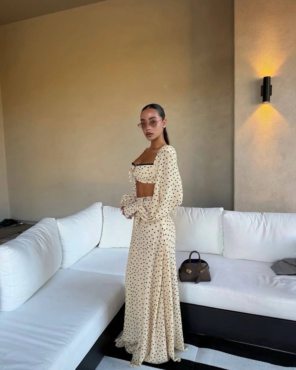 Woman in a polka dot dress standing in a room with a white couch and wall-mounted light fixture.
