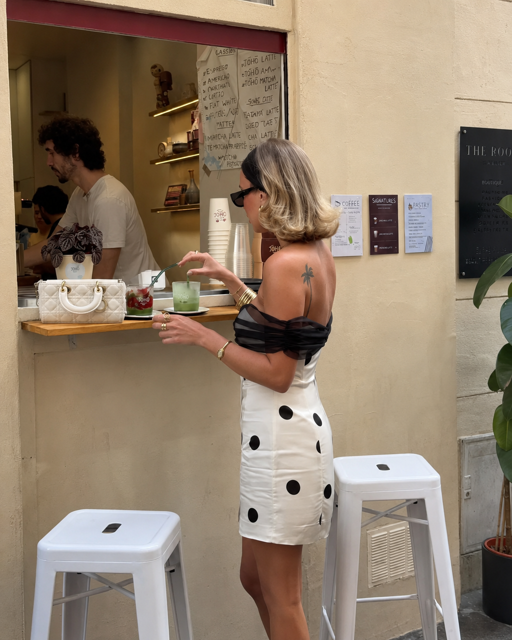 A woman in a black and white polka dot off-the-shoulder dress stands at a cafe counter, holding a drink. The background shows a cafe interior with a barista working behind the counter, white stools in the foreground, and a beige stucco wall.
