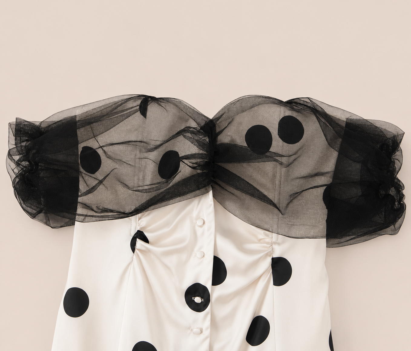A close-up of a white satin garment with large black polka dots and dramatic sheer black tulle shoulder overlays, photographed against a plain warm beige background.