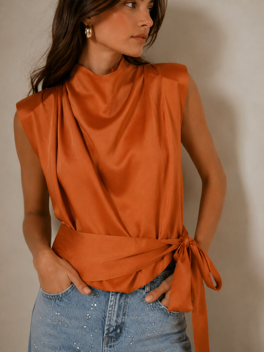 A woman wearing a sleeveless draped top in a warm burnt orange color, paired with light blue jeans, shown from the side against a neutral studio background.