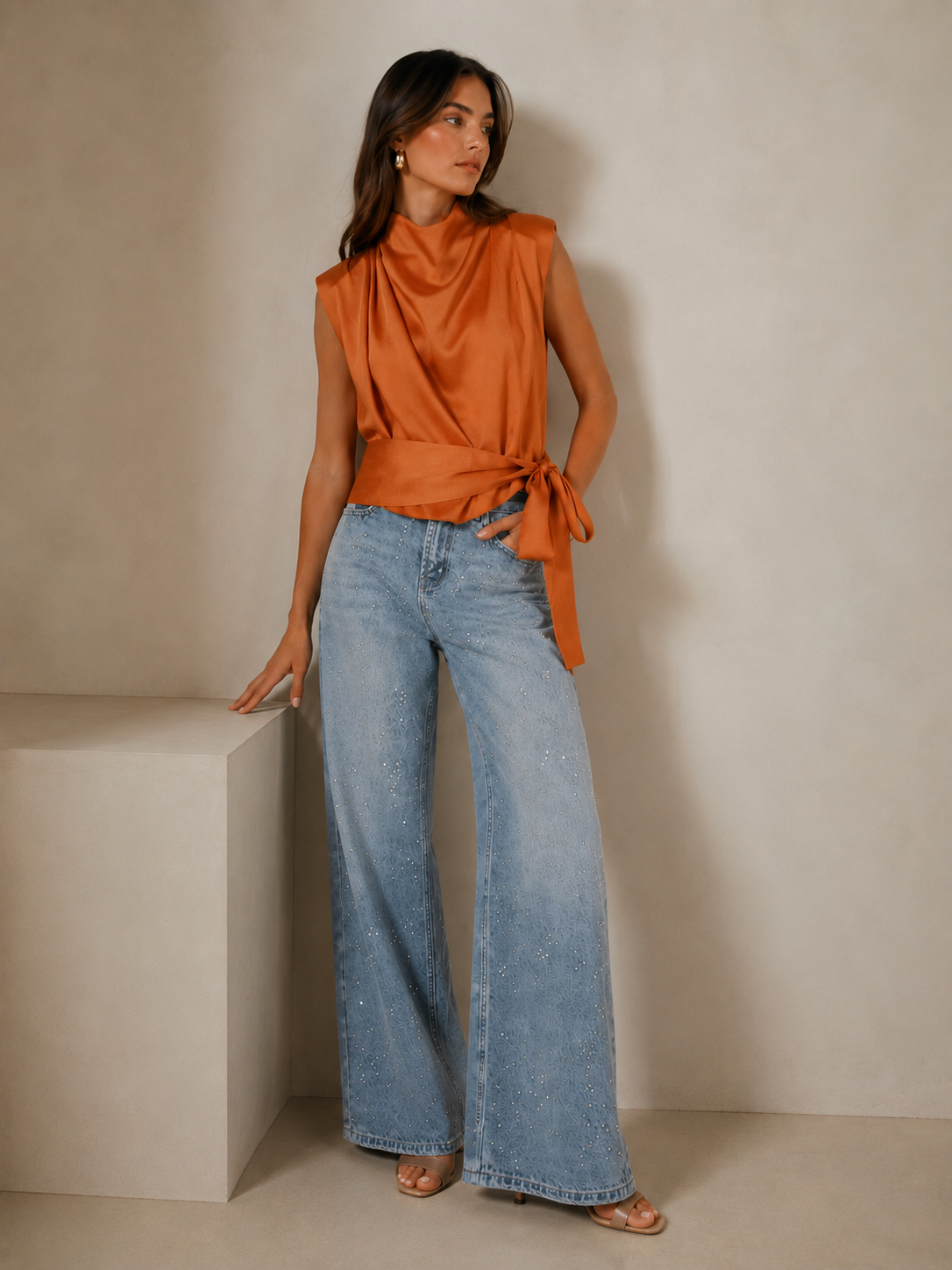 A woman models a sleeveless rust-orange satin top with a draped neckline and side tie, paired with wide-leg light-wash blue jeans, standing against a neutral beige studio backdrop.