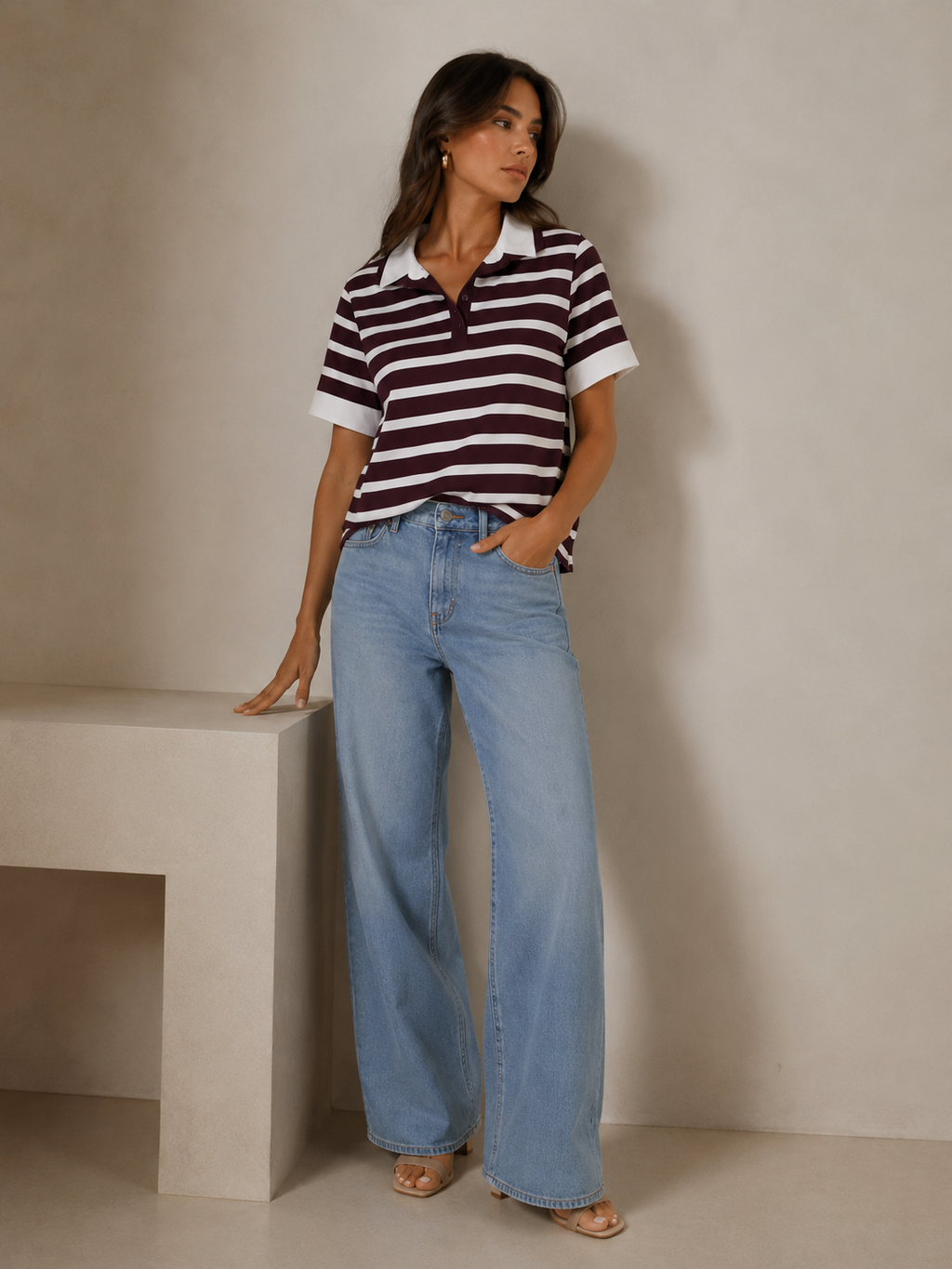 A woman stands indoors against a warm beige wall, wearing a short-sleeve polo shirt with dark burgundy and white horizontal stripes, light blue wide-leg jeans, and beige sandals.