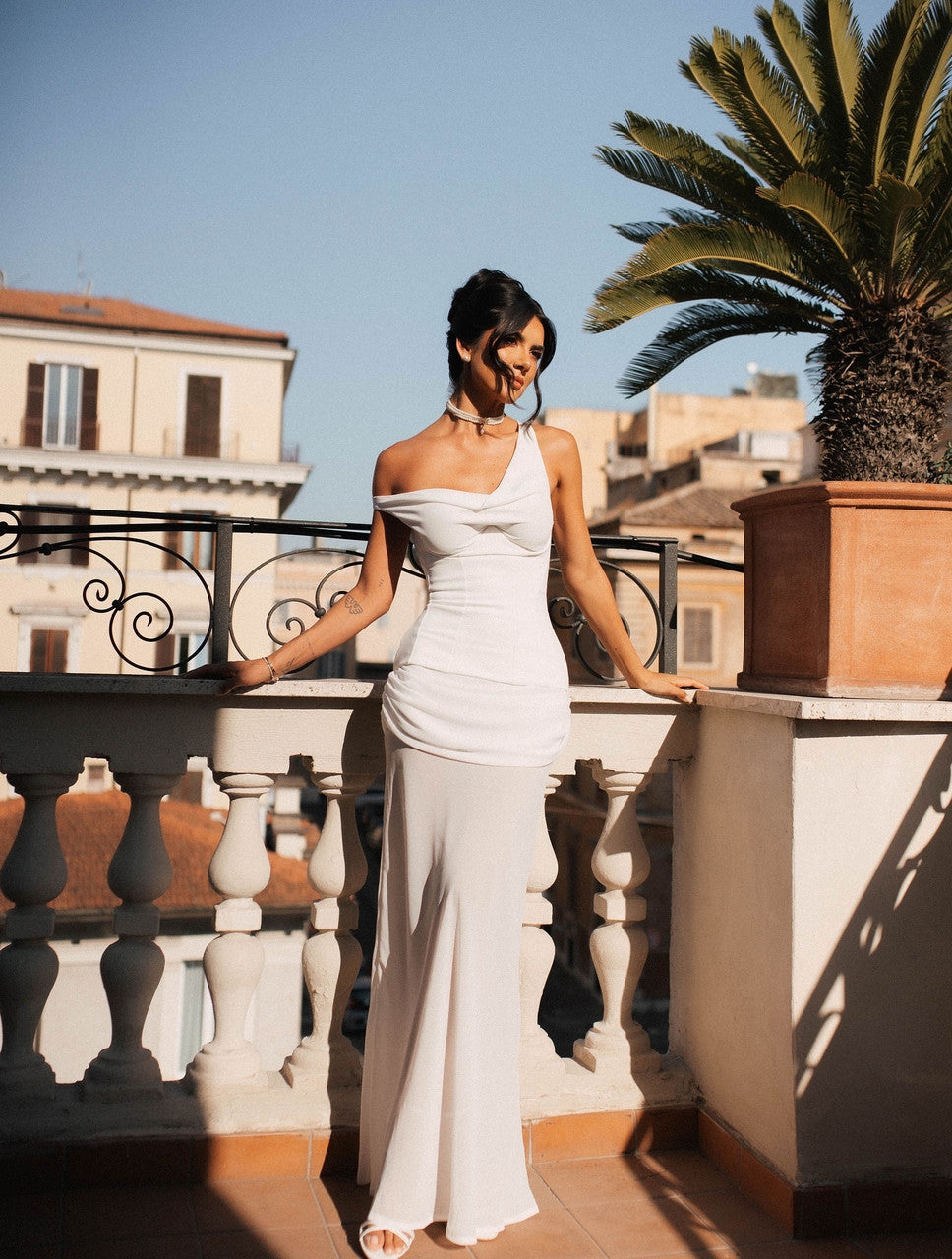 Woman in a white dress standing on a balcony with palm trees and buildings in the background